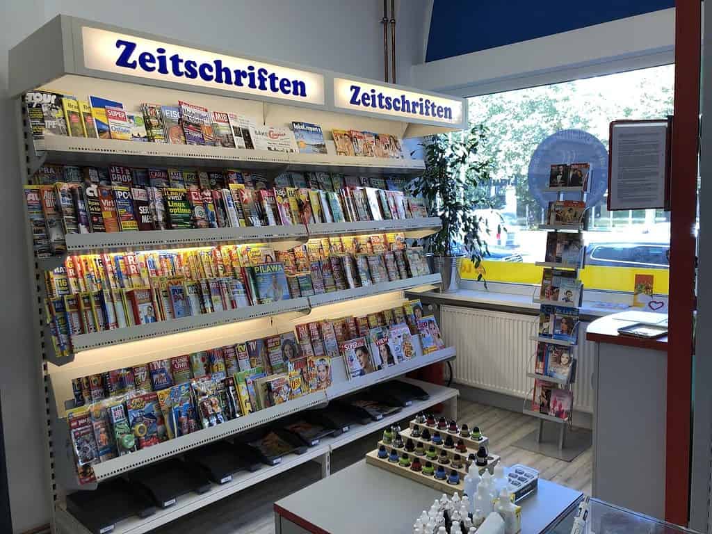 Bunte Zeitschriftenauslage mit Magazinen und Zeitschriften im Laden, sichtbar durch das Fenster, mit verschiedenen Titeln und einer Informationswand im Hintergrund.