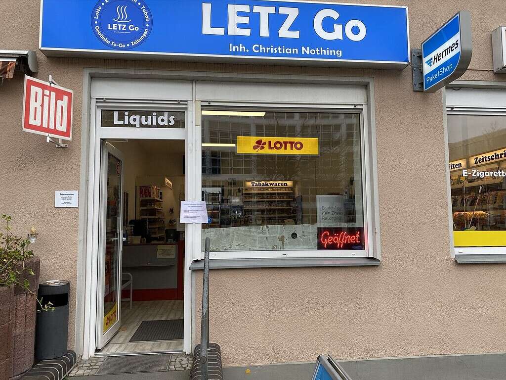 Wir haben geöffnet. 6 Gewerbeeinrichtung vom Ladeneingang von LETZ Go, mit Schaufenster, Werbung für Lotto, Tabakwaren und Liquids, Außenschild mit Logo, Sicht auf Ladeninneres.