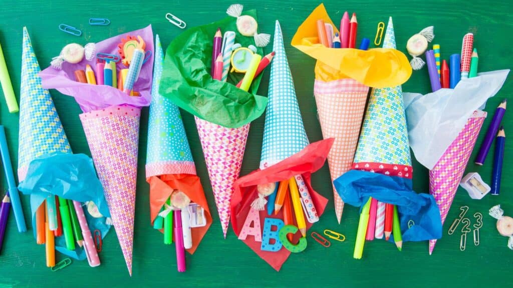 Schulanfang in Brandenburg 12 Bunte Partyhüte mit Schreibutensilien, Papier und Klammern, ideal für Kinderfeste und Schulveranstaltungen. Farbig dekorierte Hüte mit Stiften, Markern, Schleifen und Zubehör auf grünem Hintergrund.