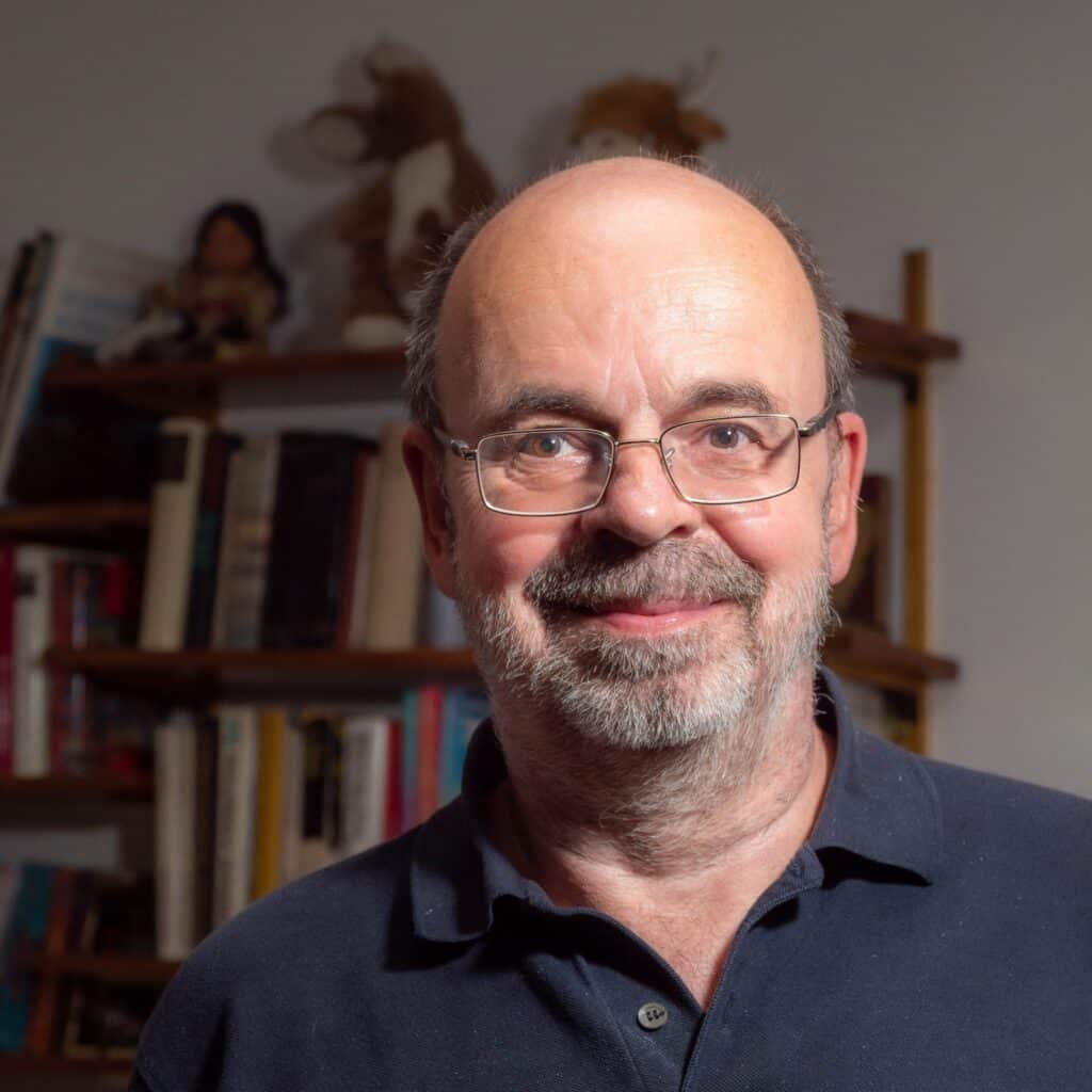 Bernd Mayer Die E-Zigarette. Fakten & Mythen 2 Mann mittleren Alters mit Brille, dunklem Polohemd, lächelnd vor Bücherregal mit Büchern und Dekorationsobjekten, freundliches Porträt, professionelle Aufnahme.