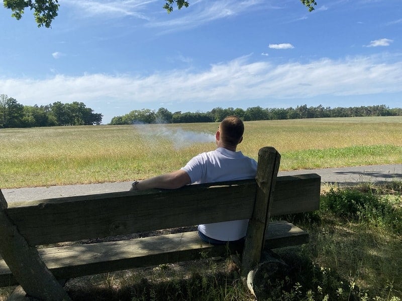 Vom Rauchen zur E-Zigarette: Eine einfache Anleitung für Anfänger 1 Entspannter Mann sitzt auf Holzbank am Feld und raucht eine Zigarette, an sonnigem Tag mit blauen Himmel und wenigen Wolken, ruhige Naturkulisse, entspannte Freizeitgestaltung.