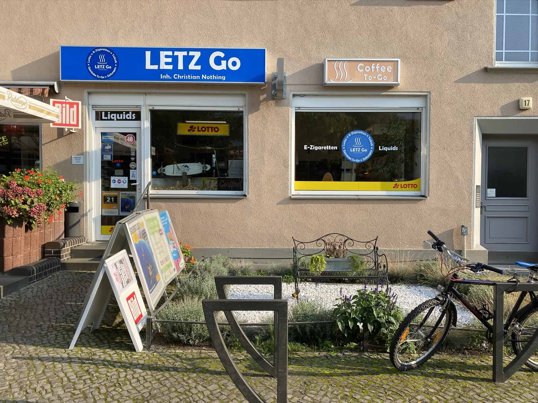 LETZ-Go-Geschäftsfront-mit-externen-Marketingtafeln-und-Fahrrad-auf-gehobenem-Gehweg, Außenansicht des Ladens, Spezialist für E-Zigaretten, Zigaretten, Liquids, Lotto-Service, Kaffee To-Go, freundliche Atmosphäre, attraktiv gestaltet.