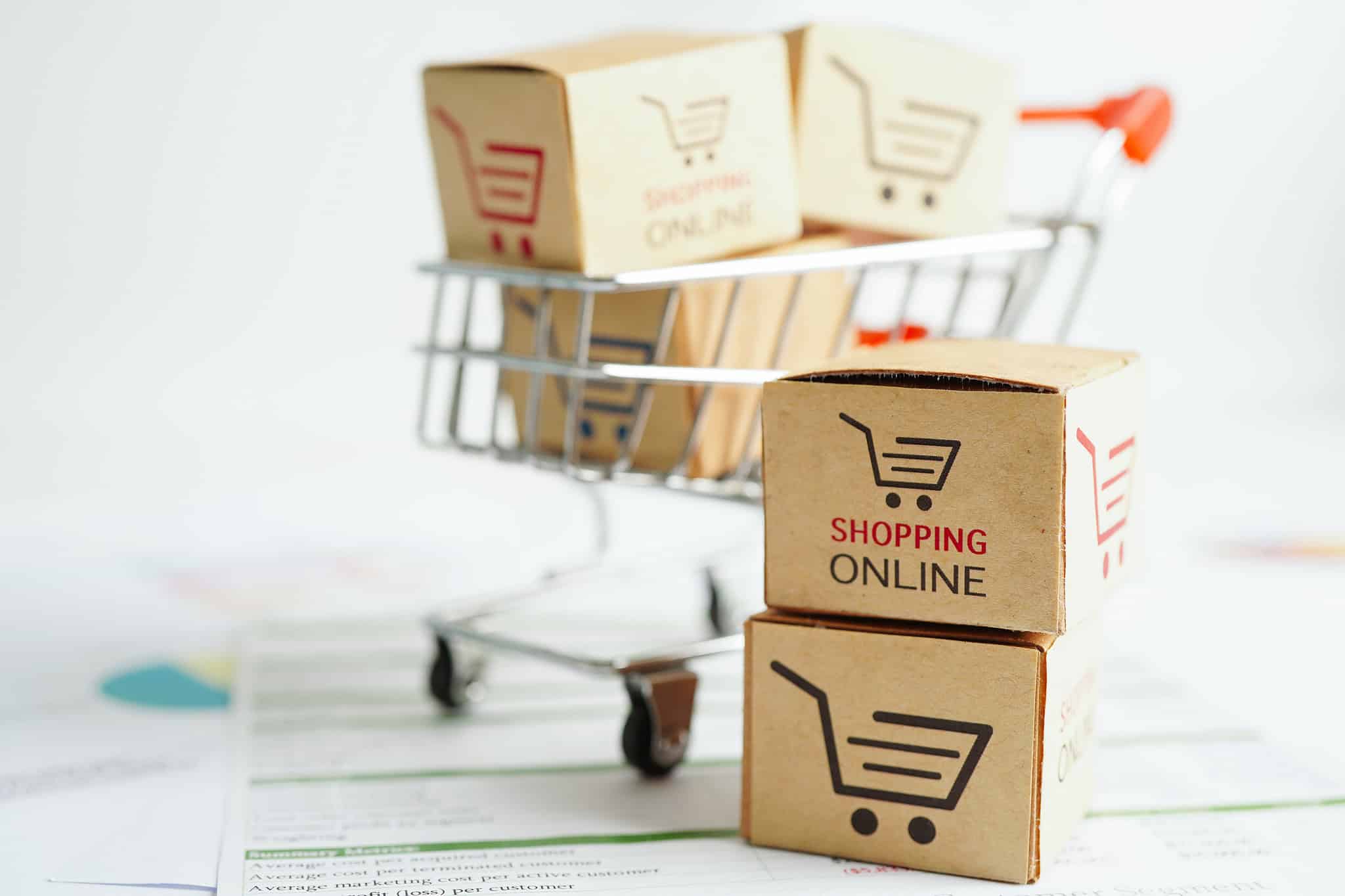 Miniaturisierte Pappkartons mit "Shopping Online" Logo auf einem kleinen Einkaufskorb, symbolisieren den Online-Einkauf.